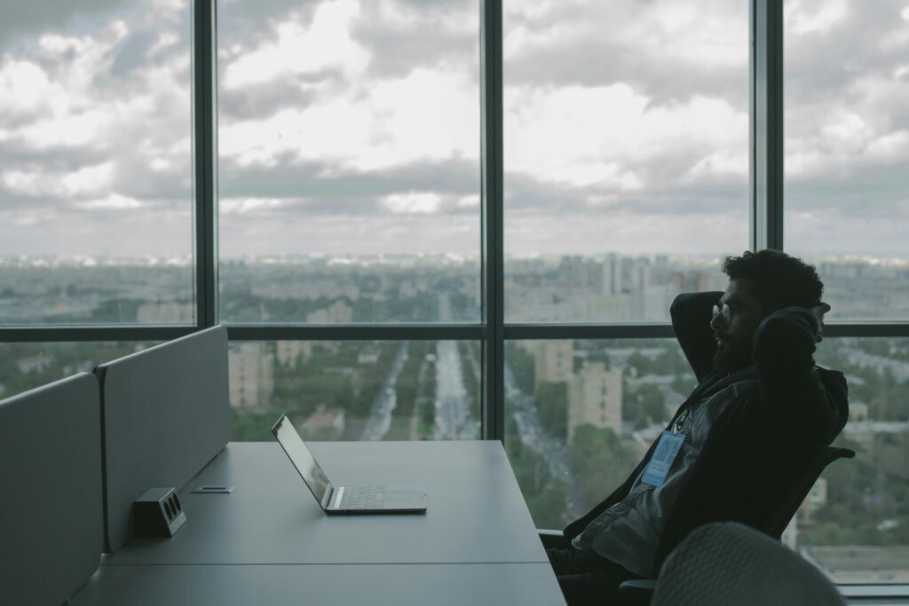 A man sits in an office with hands on head in front of a laptop, overlooking a cityscape.